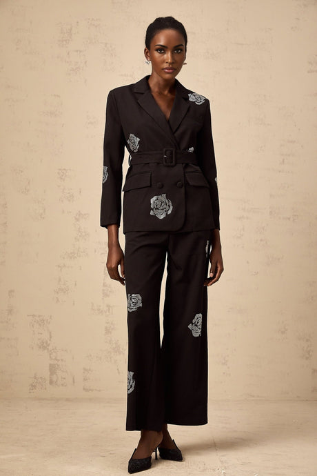 A woman wearing a black blazer and trousers set with studded rose details The blazer features a tailored fit and the trousers are slim cut Both pieces have decorative rose patterns made with small studs