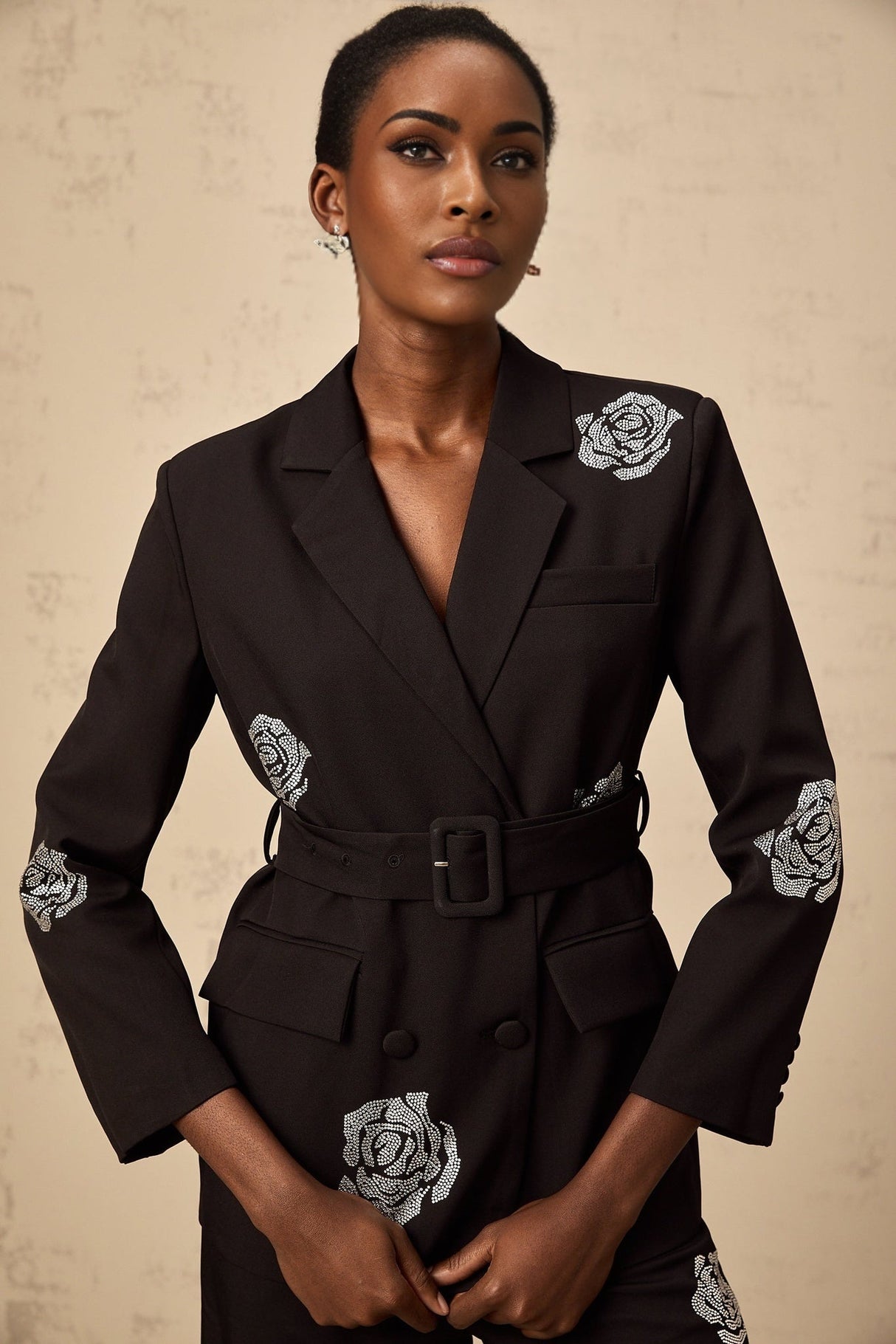 A woman wearing a black blazer and trousers set with studded rose details The blazer features a tailored fit and the trousers are slim cut The outfit is adorned with small metallic studs forming rose patterns