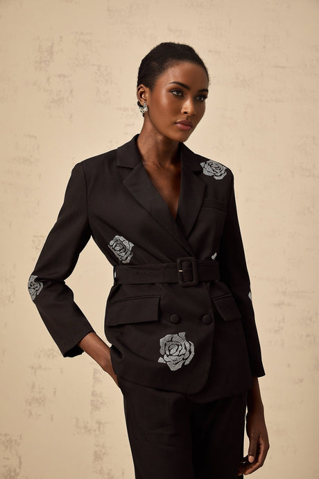 A woman wearing a black blazer and trousers set with studded rose details The blazer features a tailored fit and the trousers are slim fit Both pieces have decorative metal studs arranged in rose patterns