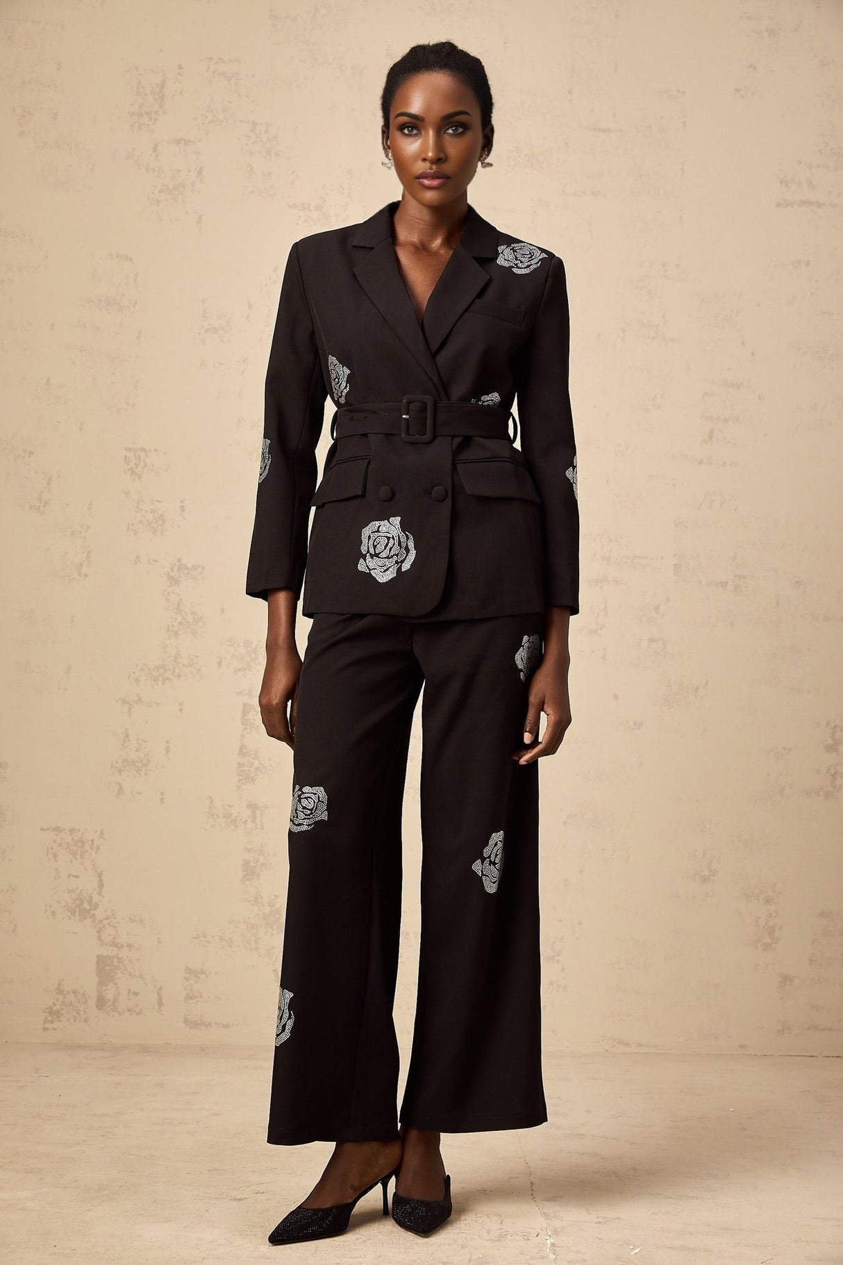 A woman wearing a black blazer and trousers set with studded rose details The blazer has a tailored fit and the trousers are sleek Both pieces feature decorative studded rose patterns throughout