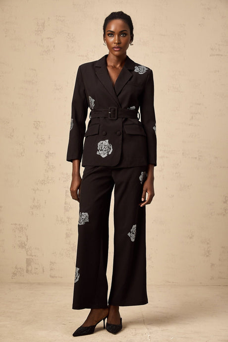 A woman wearing a black blazer and trousers set with studded rose details The blazer has a tailored fit and the trousers are sleek Both pieces feature decorative studded rose patterns throughout