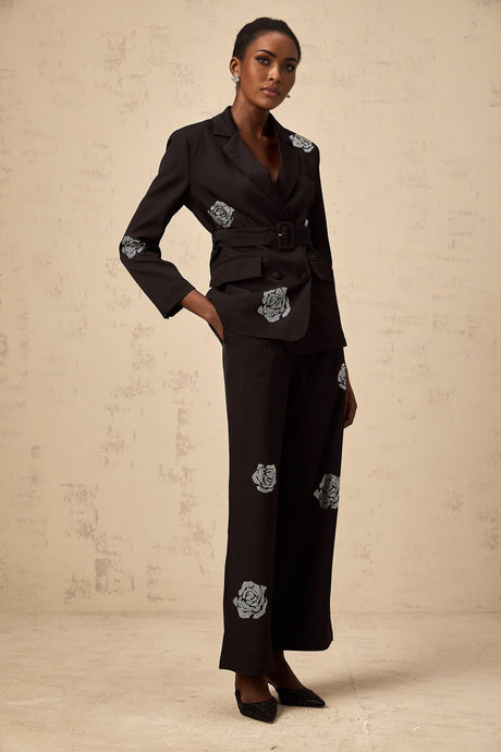 A woman wearing a black blazer and trousers set with studded rose details The blazer has a tailored fit and the trousers are slim The outfit is adorned with small metallic studs forming rose patterns