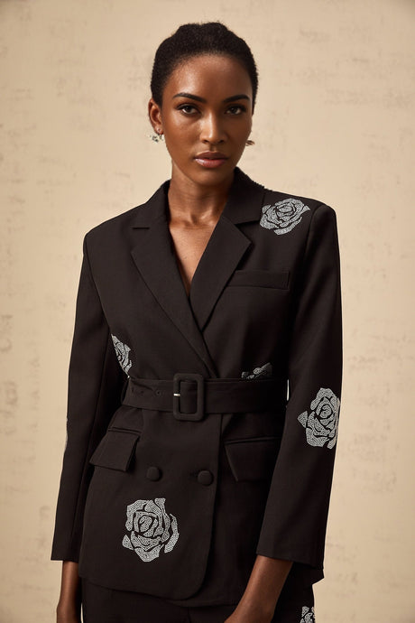 A woman wearing a black blazer with studded rose details and matching trousers standing confidently in a stylish pose