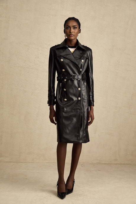 A woman wearing a black doublebreasted leather coat with a belt cinched at the waist The coat has a long length and a sleek design