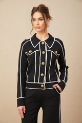 A woman wearing a black jacket and matching trousers with contrasting white piping trim along the edges standing against a plain background