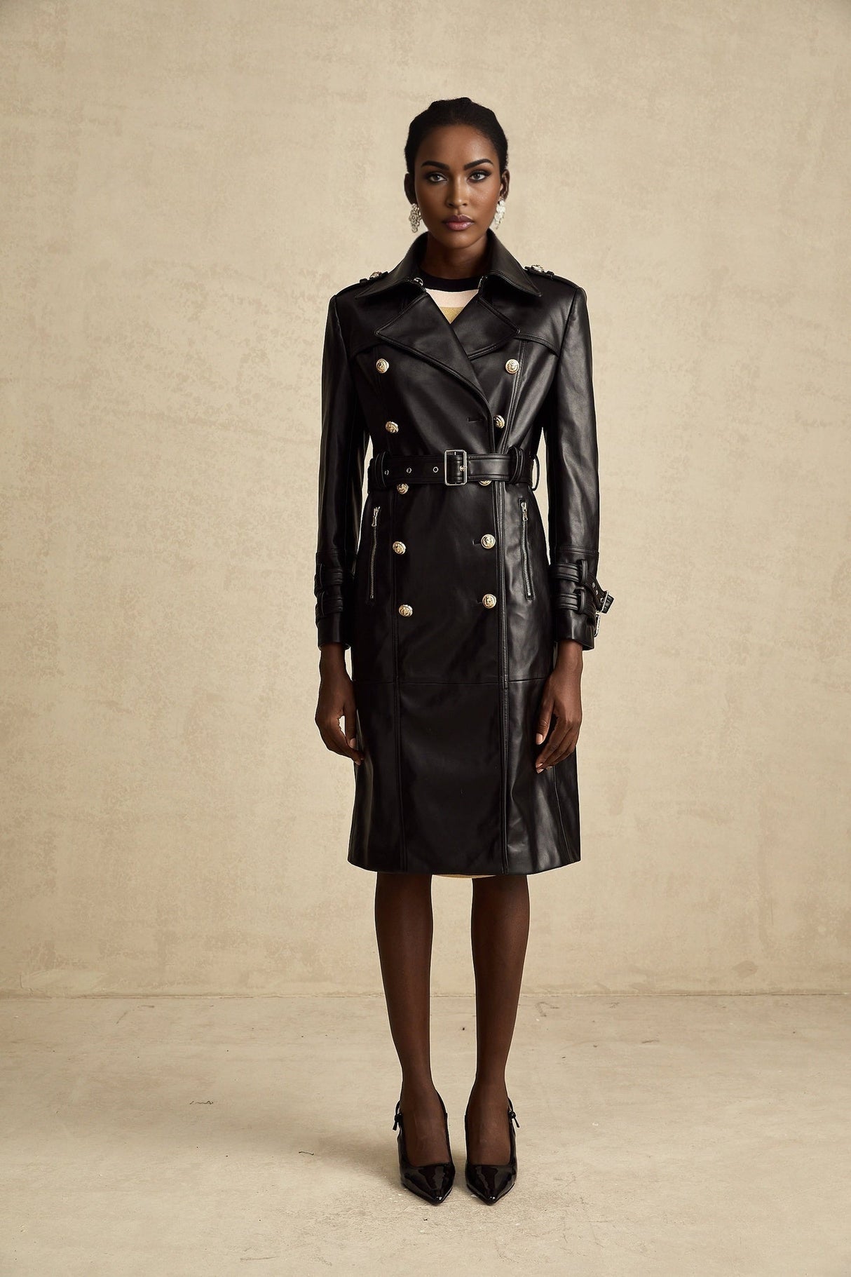 A woman wearing a black leather coat with double breasted buttons and a belt at the waist the coat has a sleek design and falls to about knee length