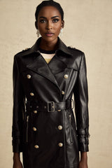 A woman wearing a black leather coat with doublebreasted front buttons and a belt at the waist The coat is long and has a classic style