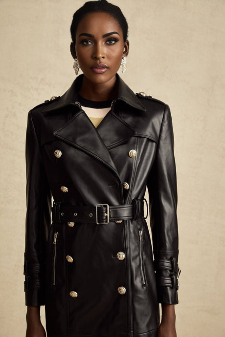 A woman wearing a black leather coat with doublebreasted front buttons and a belt at the waist The coat is long and has a classic style