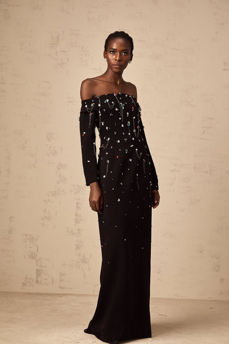 A woman wearing a black offshoulder maxi dress with crystal embellishments standing elegantly against a plain background
