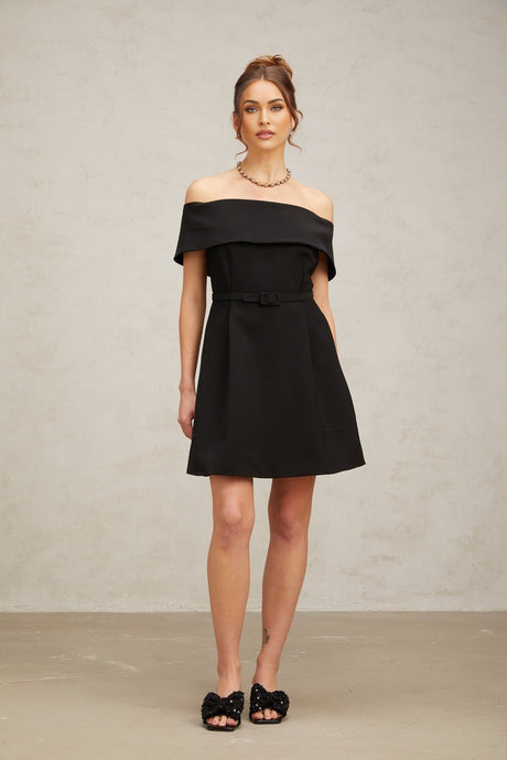 A woman wearing a black offshoulder mini dress with a belt cinching the waist standing against a plain background