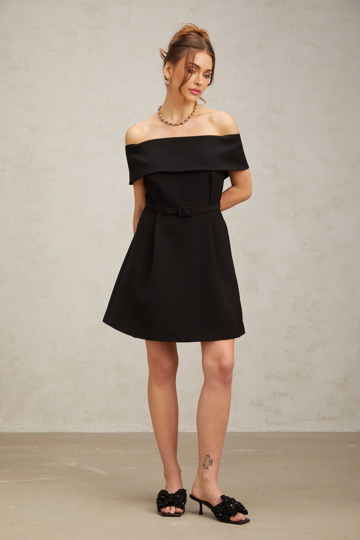A woman wearing a black offshoulder mini dress with a belt cinching the waist standing against a plain background