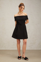 A woman wearing a black offshoulder mini dress with a belt cinching the waist standing against a plain background