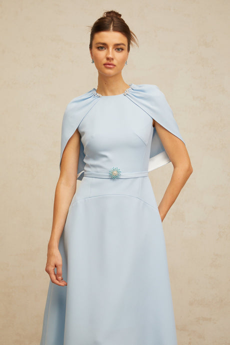 A woman wearing a blue midi dress with shawl panels and embellishments standing in a neutral background