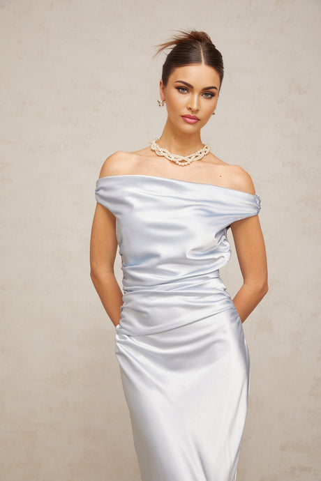 A woman wearing a blue offshoulder maxi dress with ruched satin fabric standing against a plain background