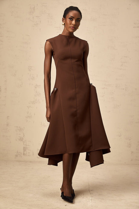 A woman wearing a brown asymmetric midi dress with one shoulder strap standing against a plain background