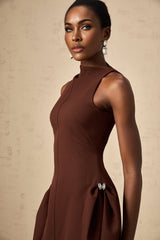A woman wearing a brown mini dress with an asymmetric neckline and puffed sleeves standing against a plain background