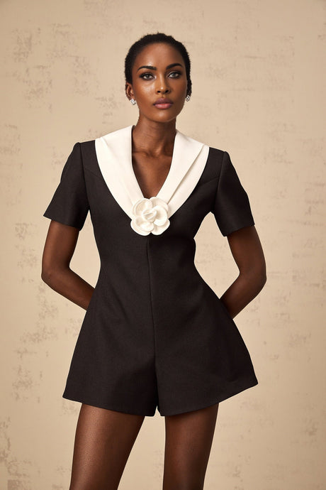 A woman wearing a colorblocked jumpsuit with floral appliqués standing in a relaxed pose against a plain background