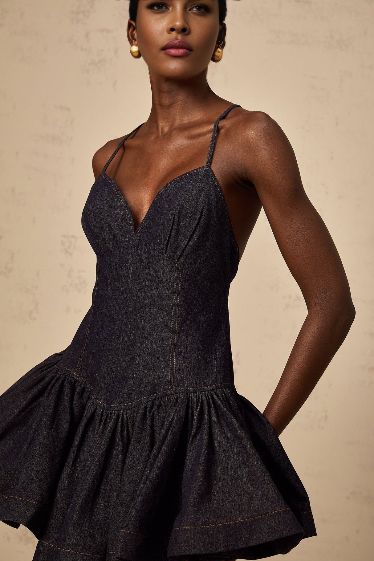 A woman wearing a dark blue denim mini dress with flounce details standing against a plain background