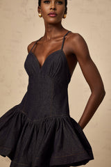 A woman wearing a dark blue denim mini dress with flounce details standing against a plain background