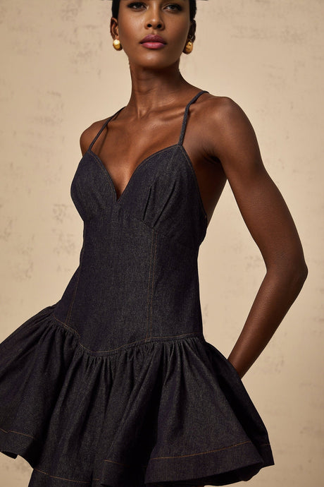 A woman wearing a dark blue denim mini dress with flounce details standing against a plain background