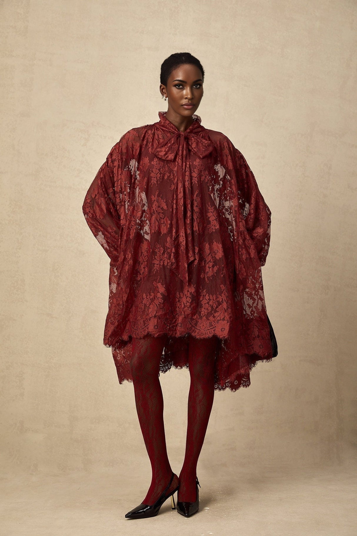 A woman wearing a flowing red oversize dress made of delicate chantilly lace standing against a plain background