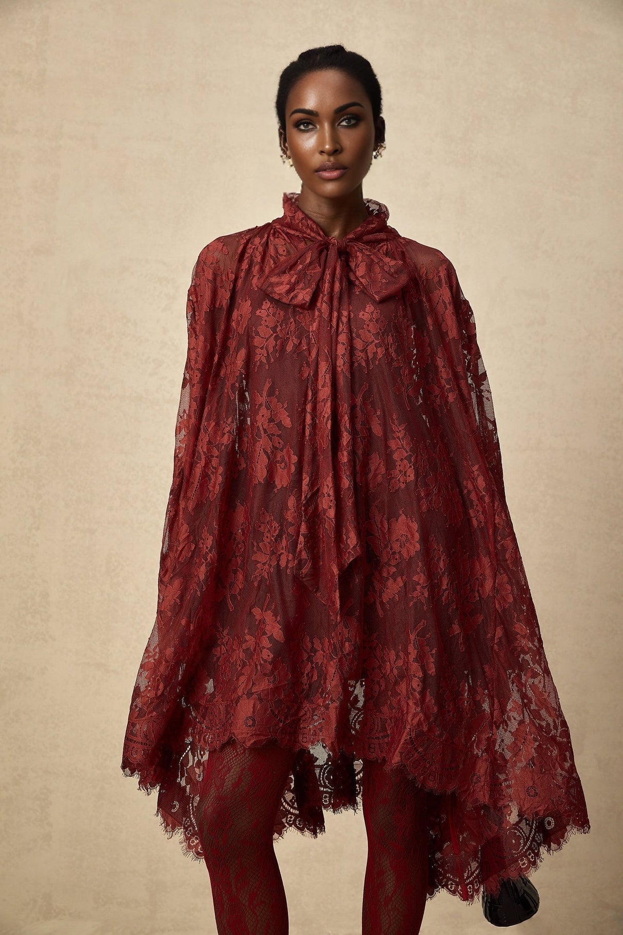 A woman wearing a flowing red oversize dress made of delicate chantilly lace standing against a plain background