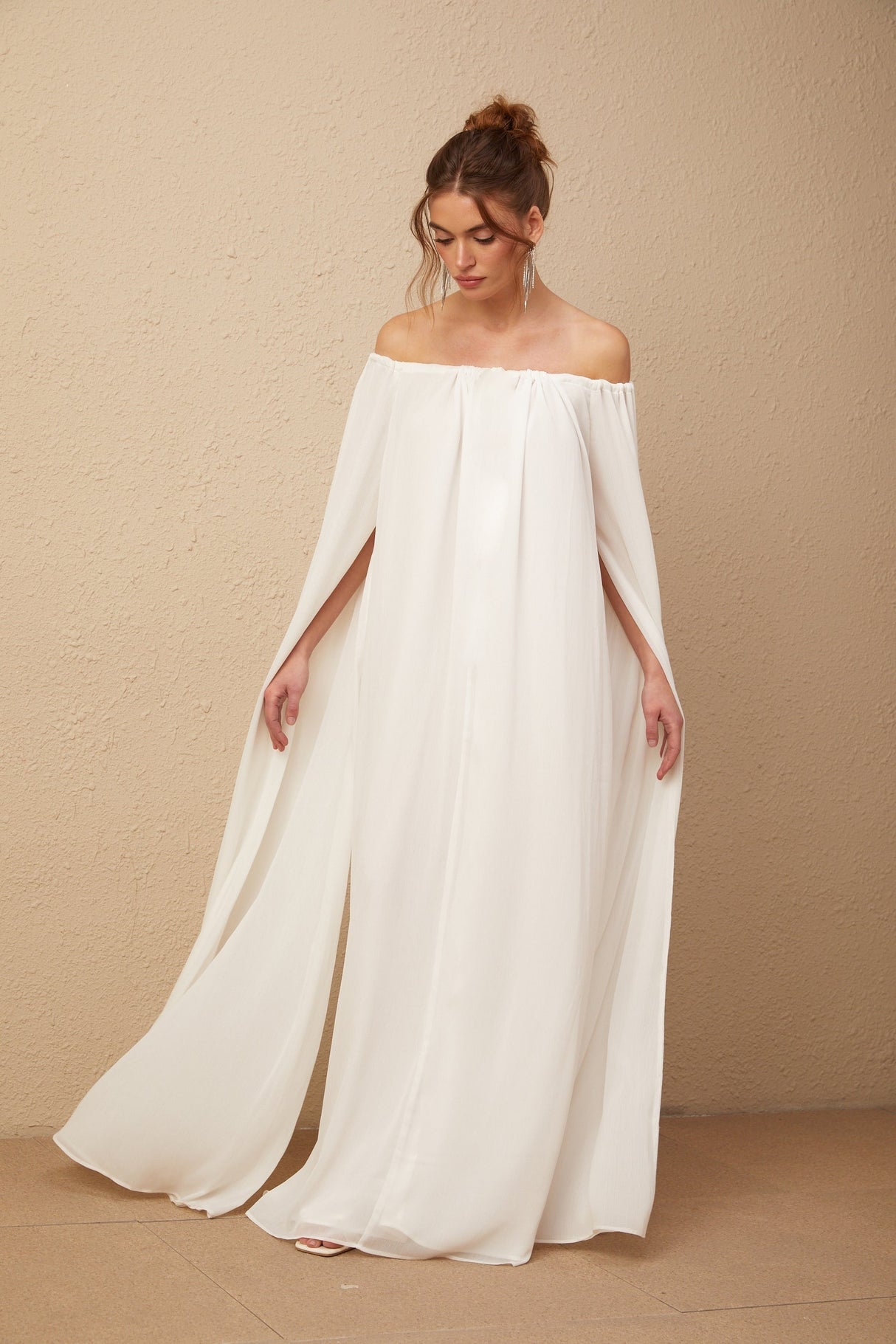 A woman wearing a flowing white tulle maxi dress with a capelike design and elegant draping details