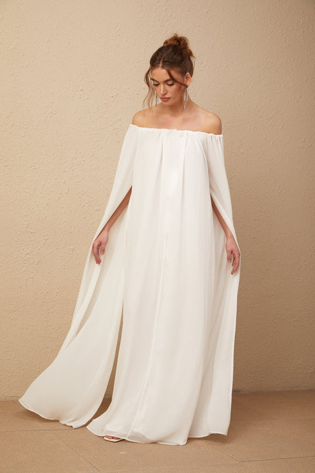 A woman wearing a flowing white tulle maxi dress with a capelike design and elegant draping details