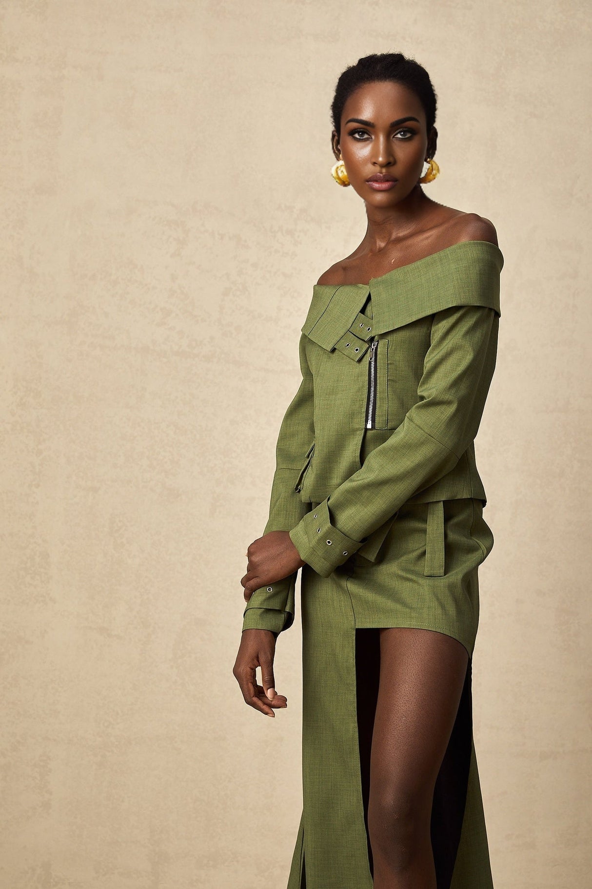 A woman wearing a green asymmetric jacket paired with a matching skirt standing against a plain background