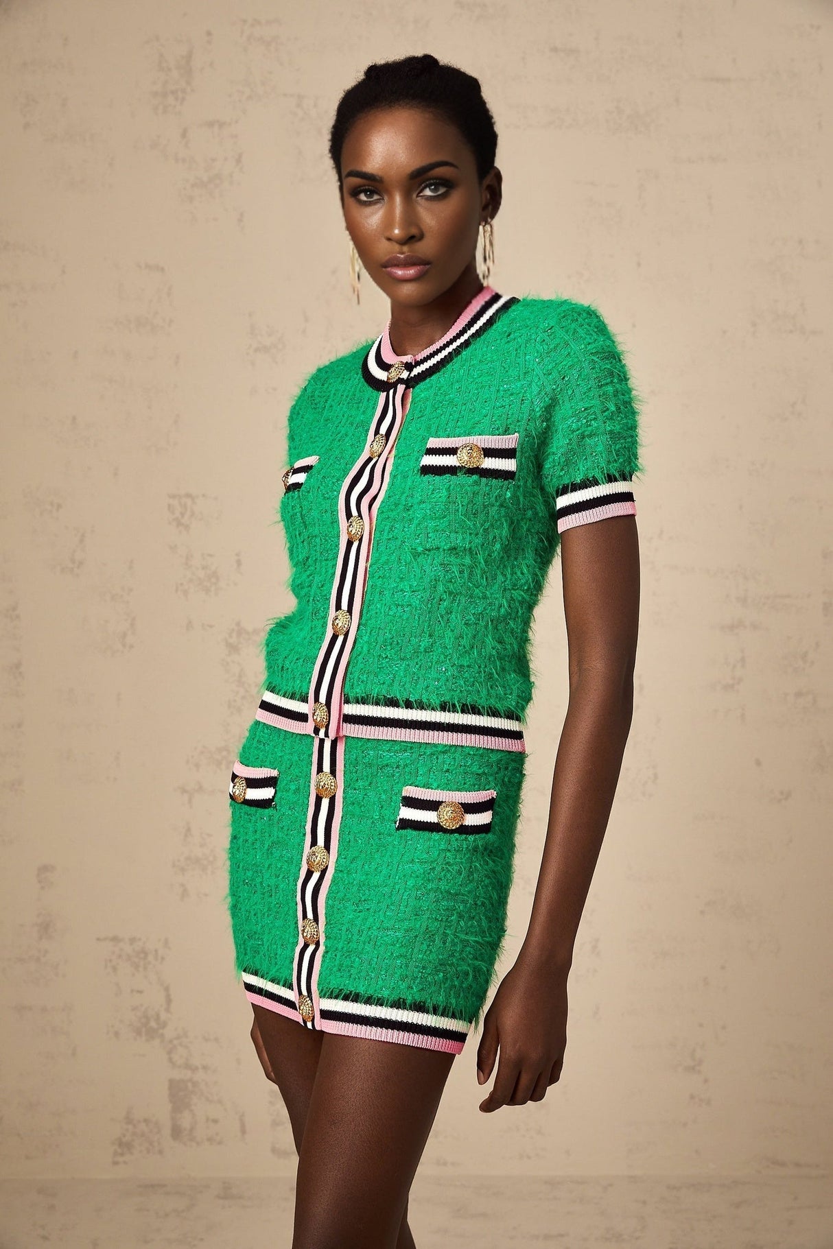A woman wearing a green knitted two piece outfit consisting of a top and skirt set The top has long sleeves and the skirt is knee length The model is standing against a plain background