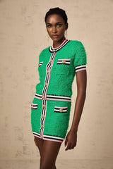 A woman wearing a green knitted two piece outfit consisting of a top and skirt set The top has long sleeves and the skirt is knee length The model is standing against a plain background