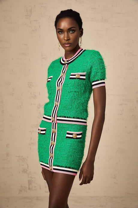 A woman wearing a green knitted two piece outfit consisting of a top and skirt set The top has long sleeves and the skirt is knee length The model is standing against a plain background