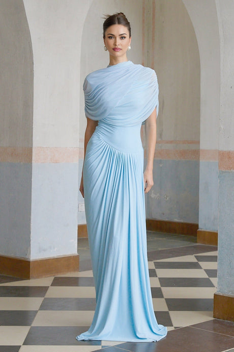 A woman wearing a light blue asymmetric pleated maxi dress with a flowing skirt and one shoulder design