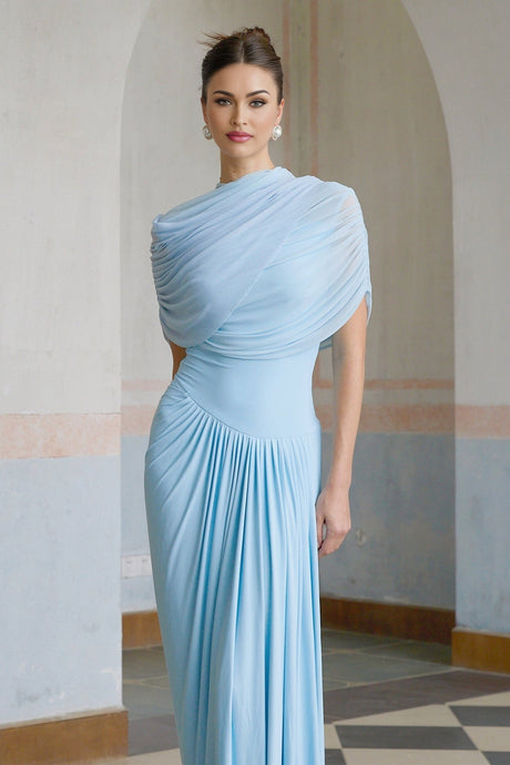 A woman wearing a light blue asymmetric pleated maxi dress with a flowing skirt and one shoulder design