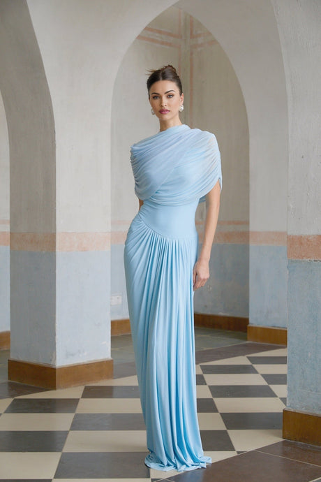 A woman wearing a light blue asymmetric pleated maxi dress with a flowy skirt and one shoulder design