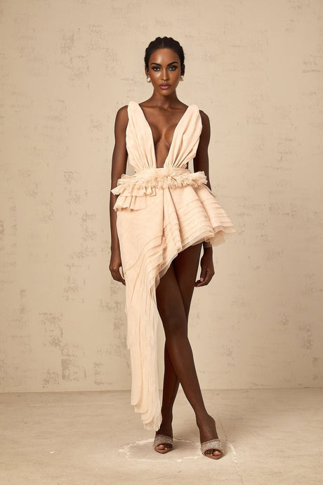 A woman wearing a lightcolored asymmetric mini dress with ribbon design details standing against a plain background