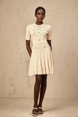 A woman wearing a lightcolored short knit dress with pleated skirt and delicate pointelle trim standing in a neutral background