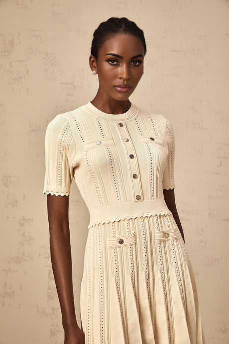 A woman wearing a lightcolored short knit dress with pleats and delicate pointelle trim details standing in a neutral background