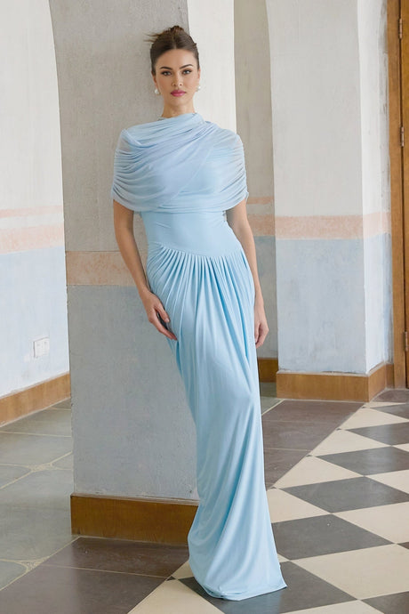 A woman wearing a long baby blue asymmetric pleated maxi dress with one shoulder strap standing against a plain background