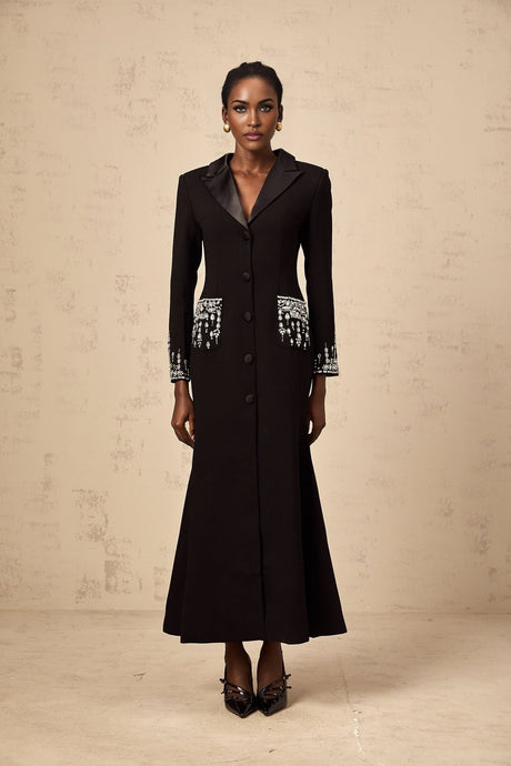 A woman wearing a long black blazer dress with embellishments standing against a plain background