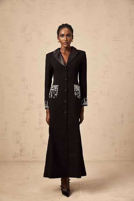 A woman wearing a long black blazer dress with embellishments standing in a neutral pose