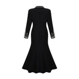 A woman wearing a long black blazer dress with embellishments standing in a relaxed pose against a plain background