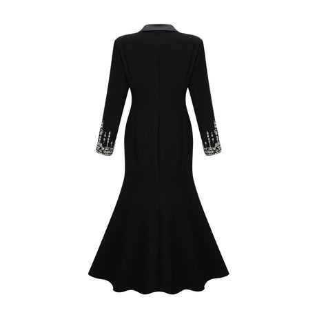 A woman wearing a long black blazer dress with embellishments standing in a relaxed pose against a plain background