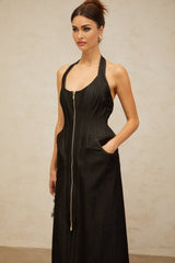 A woman wearing a long black denim maxi dress with a front slit and visible contrast stitching stands against a plain background The dress has a fitted silhouette and extends to the floor