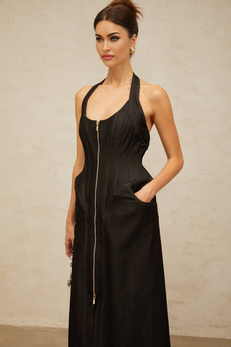 A woman wearing a long black denim maxi dress with a front slit and visible contrast stitching stands against a plain background The dress has a fitted silhouette and extends to the floor