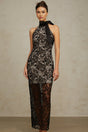 A woman wearing a long black halterneck maxi dress with floral lace pattern standing against a plain background