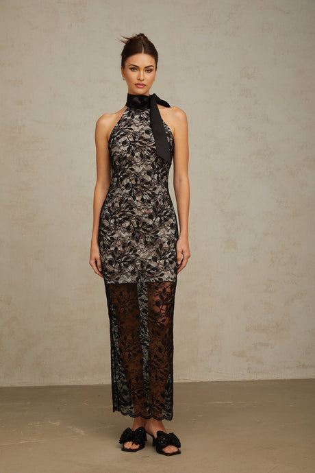A woman wearing a long black maxi dress with floral lace pattern and halter neckline standing against a plain background