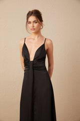A woman wearing a long black satin dress with a V neckline and a slit in the front standing against a plain background