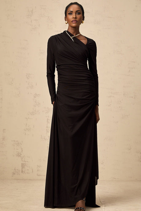 A woman wearing a long black tulle dress with an asymmetric hemline and cutout details standing against a plain background