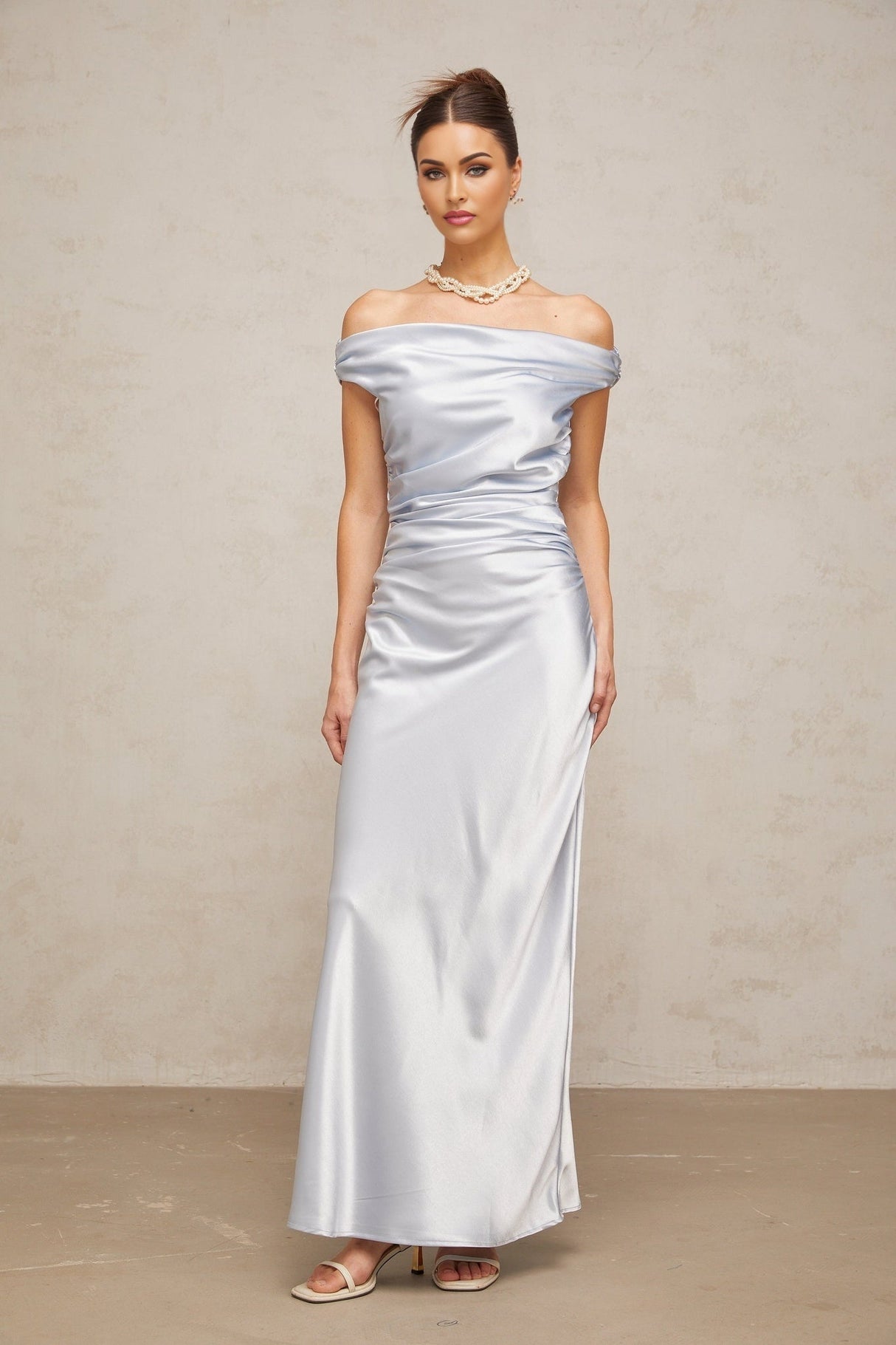 A woman wearing a long blue satin dress with offshoulder sleeves and ruched detailing standing in an elegant pose